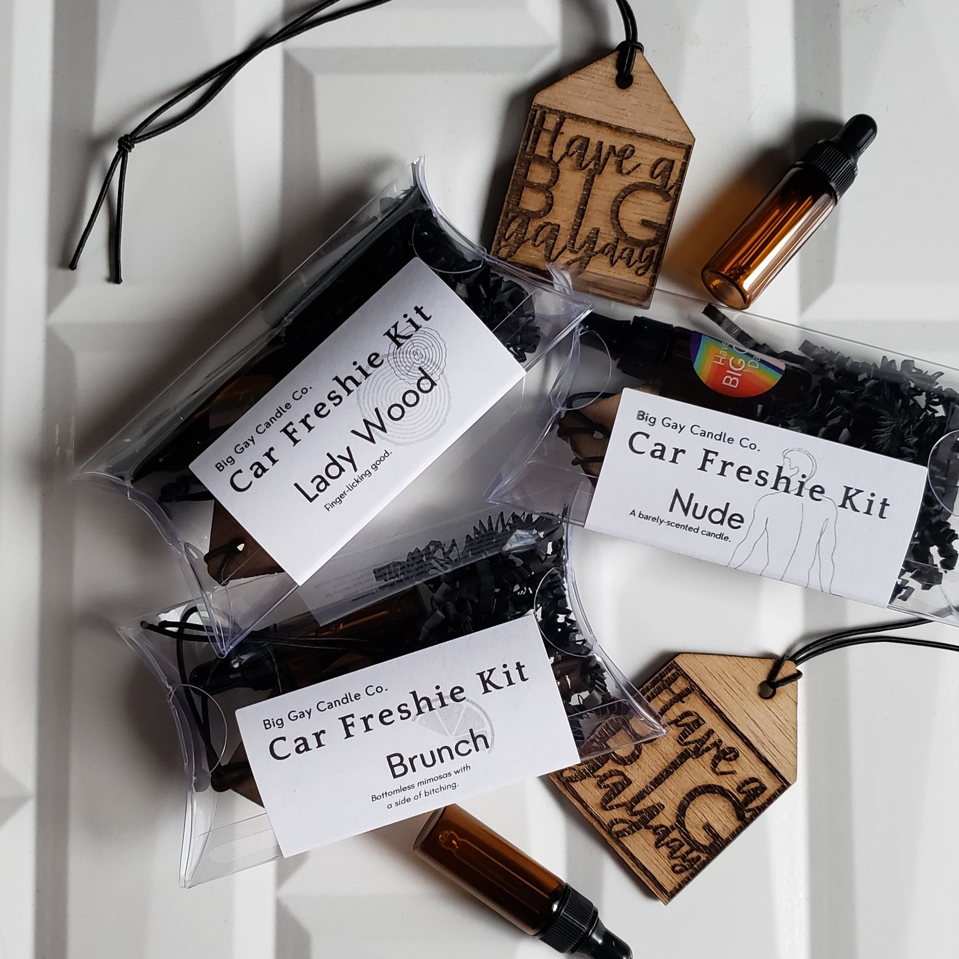 Overhead picture of a white tile background with wood, house-shaped car freshies that say Have a Big Gay Day, amber-colored glass vials of oil, and plastic pillow boxes with a black and white label with the names Lady Wood, Nude, and Brunch on top.