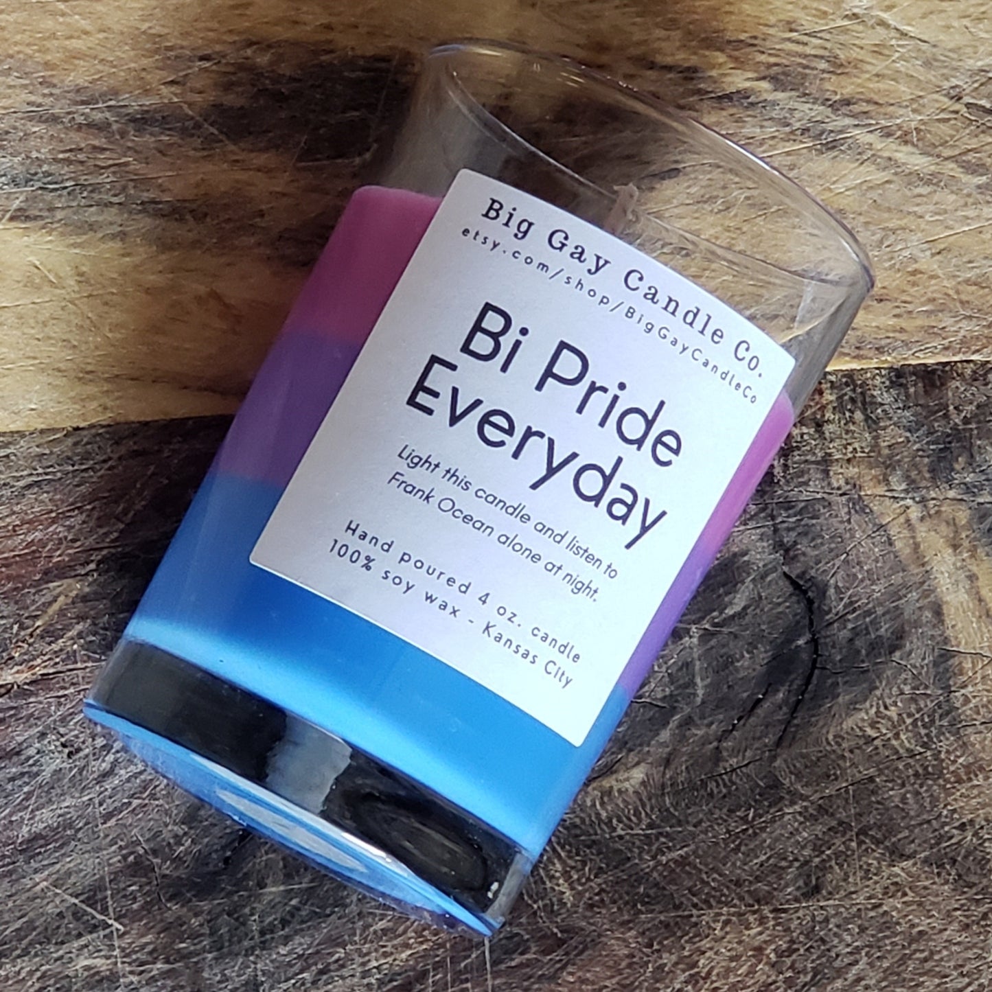 Layered candle of pink, purple, and blue wax in a glass vessel with a black and white label that says Bi Pride Everyday: Light this candle and listen to Frank Ocean alone at night, laying on a wood board.