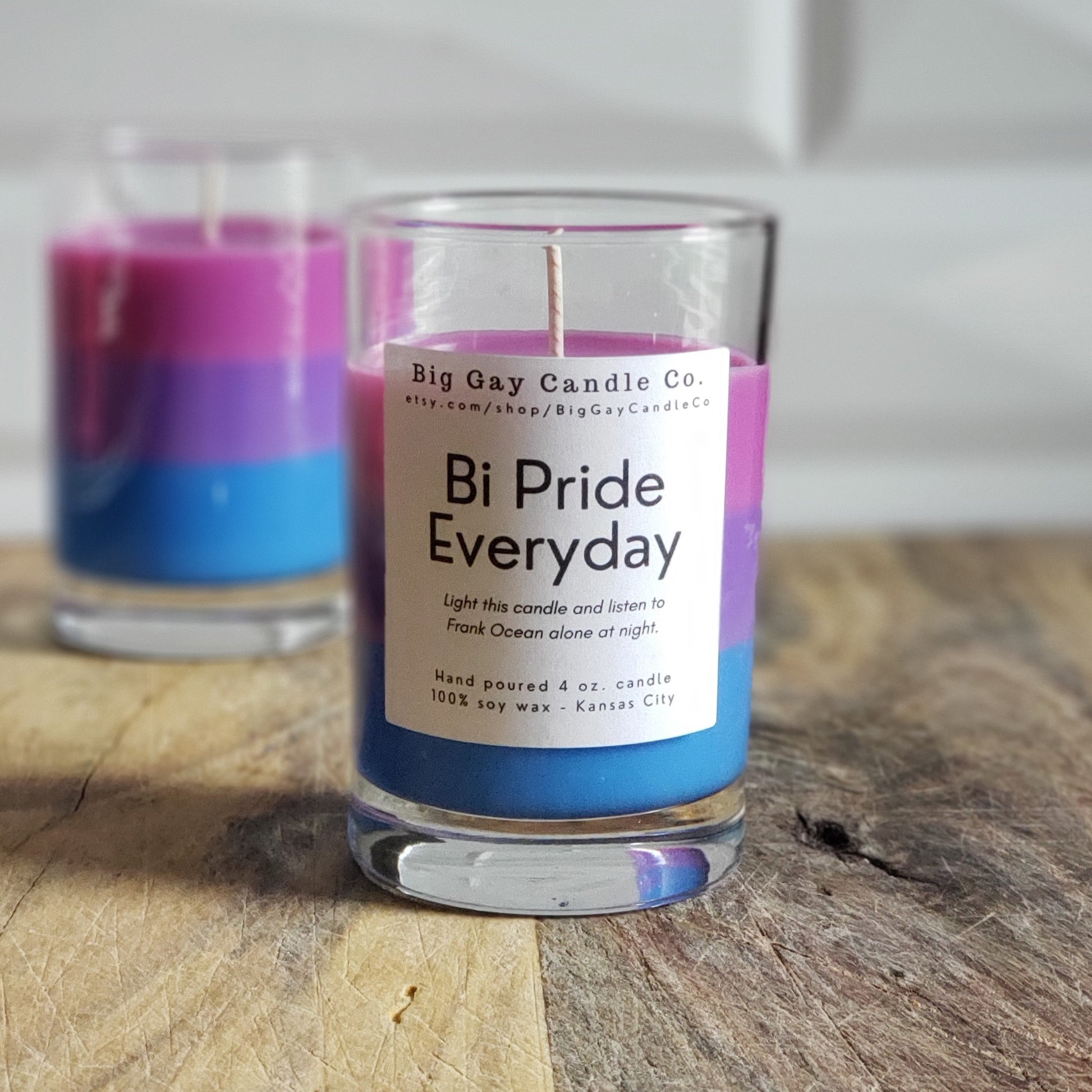 Layered candle of pink, purple, and blue wax in a glass vessel with a black and white label that says Bi Pride Everyday: Light this candle and listen to Frank Ocean alone at night. Sitting in front of another layered candle without a label in front of a white tile background and sitting on a wood board.
