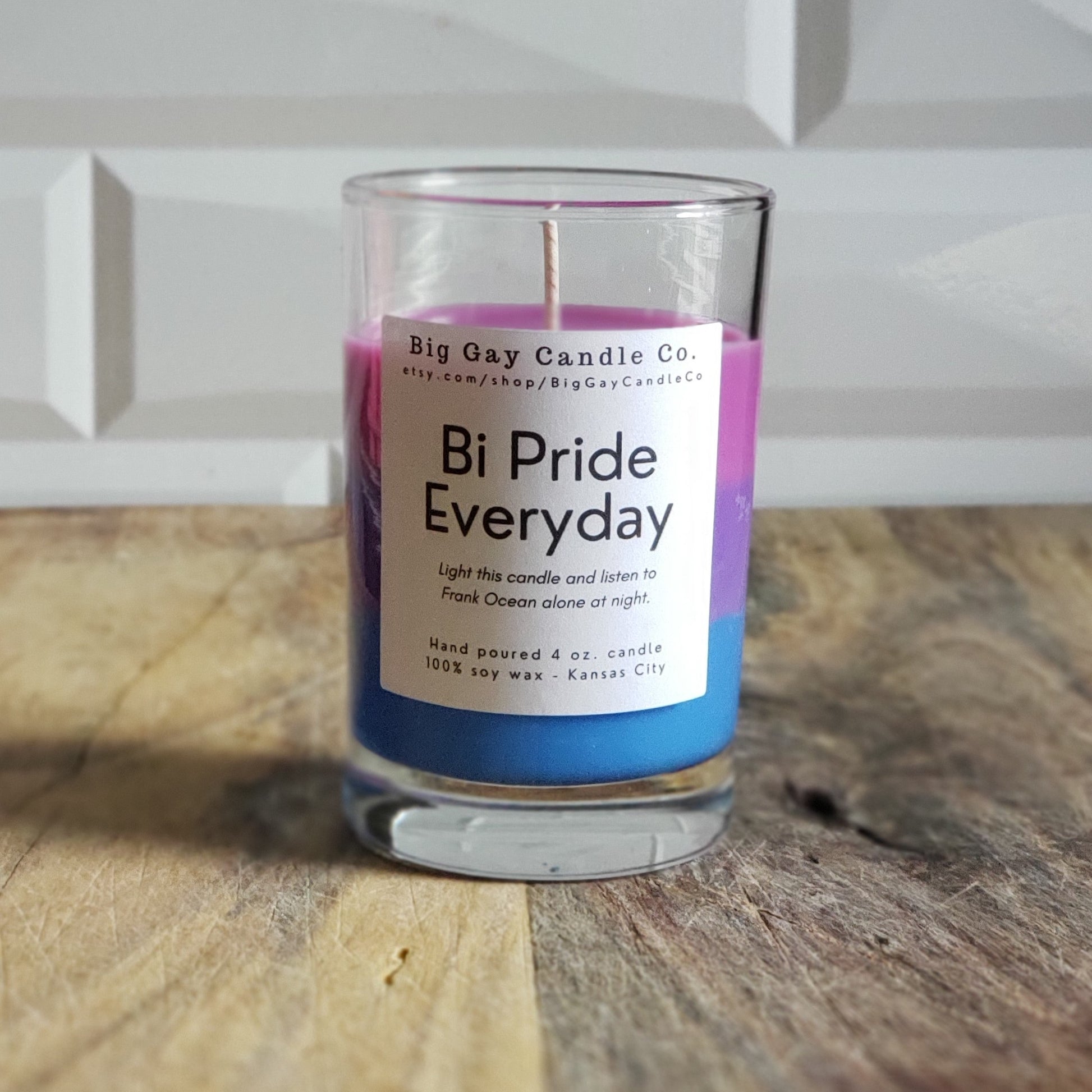 Layered candle of pink, purple, and blue wax in a glass vessel with a black and white label that says Bi Pride Everyday: Light this candle and listen to Frank Ocean alone at night. Sitting in front of a white tile background and sitting on a wood board.