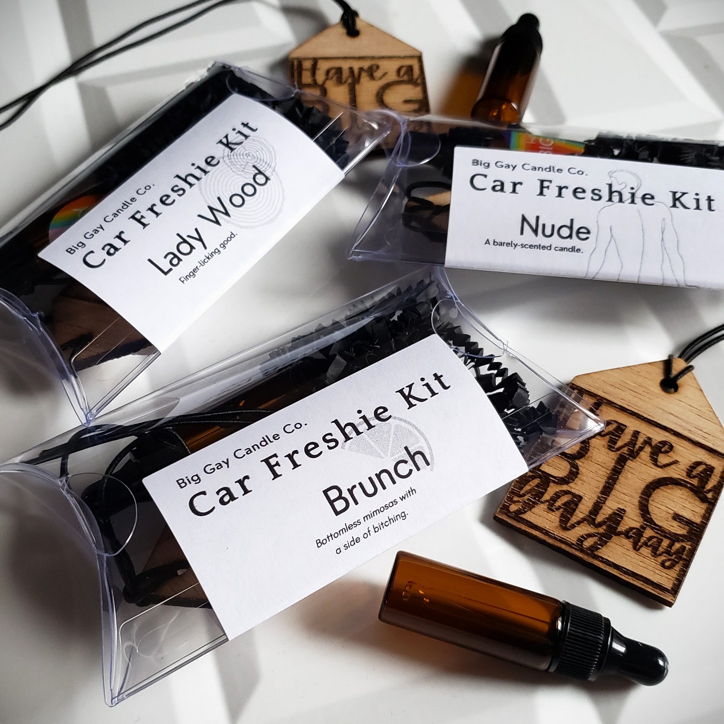 Overhead picture of a white tile background with wood, house-shaped car freshies that say Have a Big Gay Day, amber-colored glass vials of oil, and plastic pillow boxes with a black and white label with the names Lady Wood, Nude, and Brunch on top.